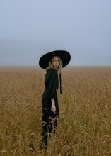 Girl In A Hat And Black Dress Posing In A Wheat Field, Foggy And Mystical Atmosphere