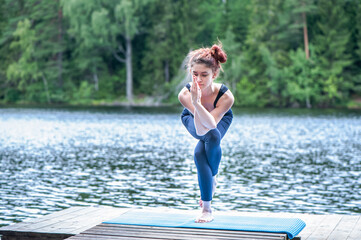 Wall Mural - Young yogi  girl  practicing yoga, standing in Eagle pose, Garudasana exercise on the lake. Concept of healthy life and natural balance