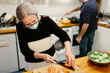 © Jimena Roquero/Stocksy - Elderly woman wearing a face mask preparing a salad in the kitchen while a man cooks in the background.