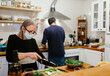 © Jimena Roquero/Stocksy - Elderly woman wearing a face mask preparing a salad in the kitchen while a man cooks in the background.