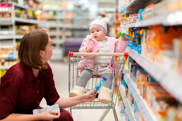 Shopping. A young mother chooses baby food with her baby sitting in a grocery cart. Close up. The concept of family shopping