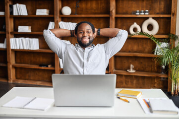 Smiling Afro-American businessman holding hands behind head sitting at office desk behind laptop. Happy black employee feeling no stress, relaxing, watching funny video after successful working