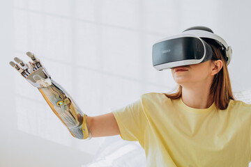 Cheerful young woman with metal bionic arm and virtual reality headset plays game near white wall in room at home closeup