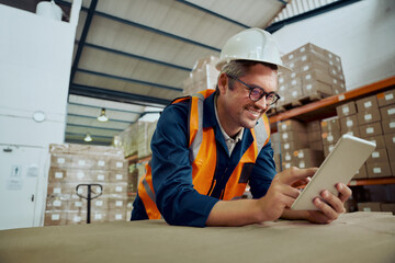 Wall Mural - Smiling factory worker leaning on table at warehouse looking at digital tablet