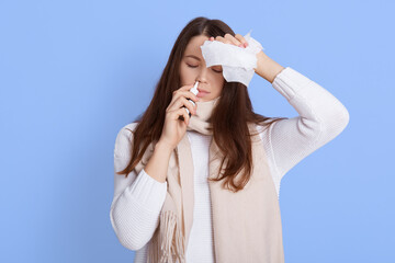  Portrait of unhappy young woman holding napkin and using nasal spray, touching her head, suffering from headache and fever, keeps eyes closed, wearing white sweater and scarf.