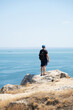 © Маргарита Гусева - A male hiker dressed in a black T-shirt, shorts, cap and backpack stands on a cliff face against the blue sky and sea on a hot sunny day