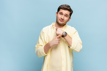 young man in yellow shirt looking at camera and pointing with finger at wristwatch on blue