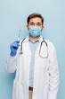 © LIGHTFIELD STUDIOS - doctor in medical mask and white coat with stethoscope holding syringe isolated on blue