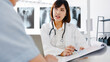 © tirachard - Young Asia female doctor in white medical uniform using clipboard is delivering great news talk discuss results or symptoms with male patient sitting at desk in health clinic or hospital office.