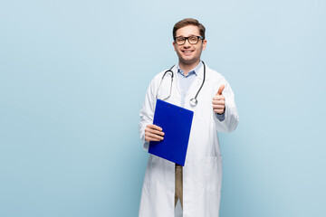 cheerful doctor in glasses and white coat pointing with finger at clipboard and showing thumb up on blue