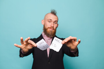 Bearded european businessman in dark suit isolated on turquoise background holding paper with you are fired tears up the paper with smile