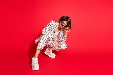 Sticker - Full body photo of young attractive business woman sit hand touch sunglass isolated over red color background