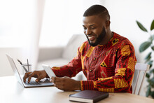 Cheerful Black Man In Traditional African Clothes Using Smartphone And Laptop