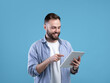 © Prostock-studio - Joyful bearded man using tablet computer for online work or studies on blue studio background
