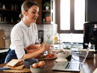 Young woman cooking in the kitchen. Beautiful woman following recipe on laptop and preparing delicious food.