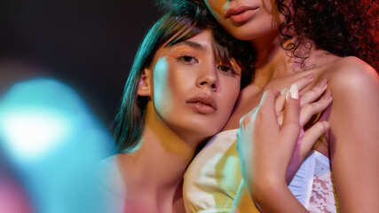 Wall Mural - Close up portrait of two artistic young female models with professional art makeup hugging, posing together in neon light isolated over black background