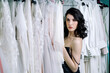 © Alex Li - Beautiful girl in black dress, hiding behind white dresses hanging on a hanger. Sensual look past the camera.