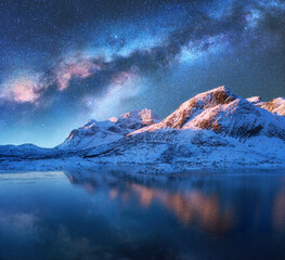 Wall Mural - Milky Way above frozen sea coast and snow covered mountains in winter at night in Lofoten Islands, Norway. Arctic landscape with blue starry sky reflected in water, snowy rocks, milky way. Space