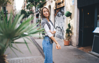 Wall Mural - Pensive female blogger with modern cellphone device in hand standing at urban street and thoughtful looking away during sightseeing time on vacations, pondering woman in trendy clothing thinking