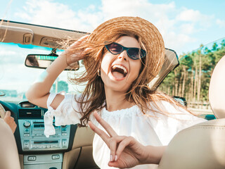 Portrait of young beautiful and smiling hipster female in convertible car. Sexy carefree woman driving cabriolet. Positive model riding and having fun in sunglasses at sunset. Enjoying summer days