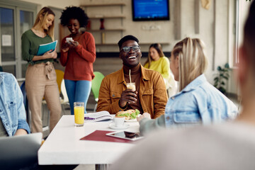 Wall Mural - Cheerful black student talking to female friend during lunch break in cafeteria.