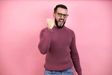 Wall Mural - Handsome man wearing glasses and casual clothes over pink background angry and mad raising fist frustrated and furious while shouting with anger. Rage and aggressive concept.