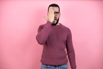 Wall Mural - Handsome man wearing glasses and casual clothes over pink background covering one eye with hand, confident smile on face and surprise emotion.