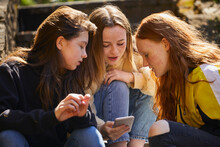 Three Teenage Girls Sitting Outdoors, Checking Their Mobile Phones.