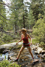 A Woman Wearing Shorts And A Tee Shirt, Sunglasses And A Straw Hat Standing On Rocks In A Stream In The Forest.