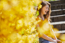 Teenage Girl Sitting Outdoors On Steps, Yellow Forsythia In Foreground.
