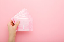Young Adult Woman Hand Holding Packs Of Sanitary Towel On Light Pink Table Background. Pastel Color. Closeup. Empty Place For Text. Top Down View.