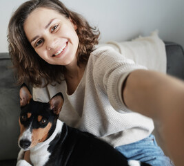 Wall Mural - Pretty curly happy young woman smiling taking selfie on smartphone with dog