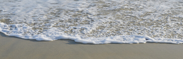 Écume blanche d'une vague sur le sable fin d'une plage ensoleillée, vue de face, formant une ligne horizontale, format bannière