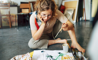 Wall Mural - Creative female artist sitting on the floor in the art studio and painting on paper with a paintbrush. A woman student painter in eyeglasses painting with watercolors in her workshop.