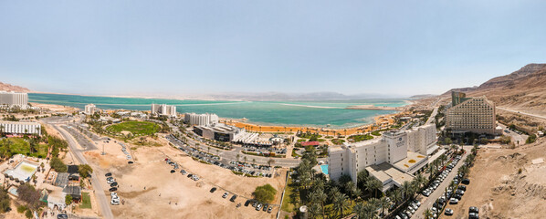 Aerial panoramic view from a drone of beach on Ein Bokek embankment on coast of Dead Sea, tourist hotels and car parks, the sea itself and the mountains of Jordan visible in the distance, in Israel