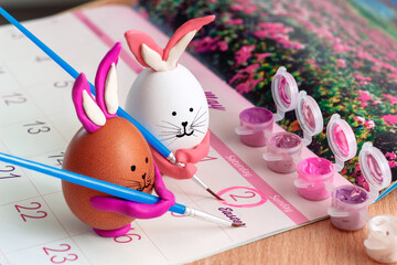 Close up of a white and brown eggs in the form of a bunny rabbit mark the date of Easter on May 2, 2021 with a pink paint brush in an open calendar. Children's art. Creative easter concept