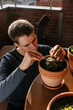 © Edgar - Hombre caucásico joven en la terraza de su casa cuidando sus plantas agachado y concentrado con varias macetas encima de la mesa