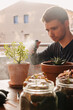 © Edgar - Hombre joven caucásico rociando una planta con agua encima de la mesa de la terraza de su casa. Cuidando plantas