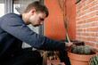 © Edgar - Hombre joven caucásico concentrado sentado en el suelo de su terraza cuidando las plantes que tiene en las macetas. Añadiendo tierra para las plantas
