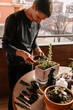 © Edgar - Hombre caucásico joven en la terraza de su casa cuidando sus plantas y suculentas con varias plantas encima de la mesa y material para cuidarlas Poniendo tierra a las plantas