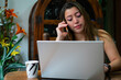 © Diego - Young latina woman using a laptop and making a phone call at home while drinking coffee. homeoffice concept