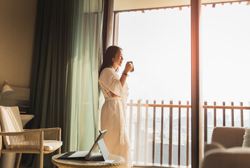 Happy businesswoman in dressing gown stands near the window holding cup of coffee.