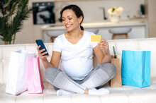 Happy Mixed-race Adult Pregnant Woman In A White T-shirt Sit On The Couch, Holds A Credit Card And A Smartphone In Hand, Makes Purchases Online, Buys Clothes And Toys For The Future Baby, Packages