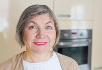 Close up portrait of happy senior woman with gray hair smiling is looking at the camera in her kitchen.