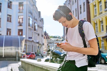 Wall Mural - Handsome guy teenager with backpack headphones looking in screen of smartphone