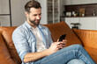 © Vadim Pastuh - Attractive millennial young bearded man with a pleasant smile chilling and sitting on the couch, chatting with friend in social media on mobile phone, dating, flirting, spending leisure time online