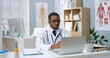 © VAKSMANV - Portrait of young handsome African American male doctor in white coat videochatting on laptop sitting in clinic in cabinet having online video consultation with patient. Healthcare, disease treatment