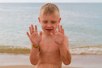 Funny caucasian boy sits on the beach by the sea in summer and laughs and shows his hands in the sand