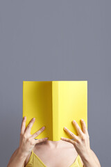 Young woman with her face hidden behind an open yellow book that she is reading