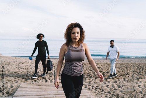 Half length portrait of curly female fitness coach with perfect figure walking at seashore and looking at camera during workout meeting with blurred male colleagues, fit girl with slim body posing